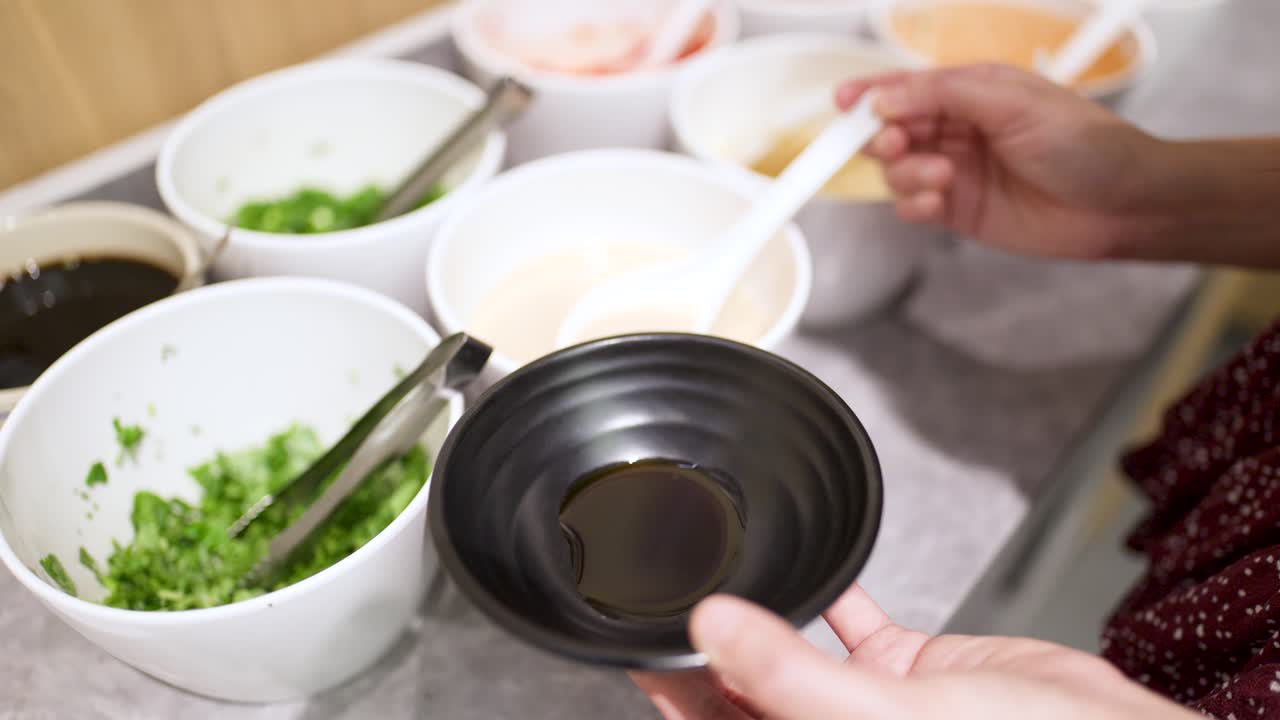 Hand swirls creamy sauce into black bowl at self-serve hotpot buffet topping station, overhead view