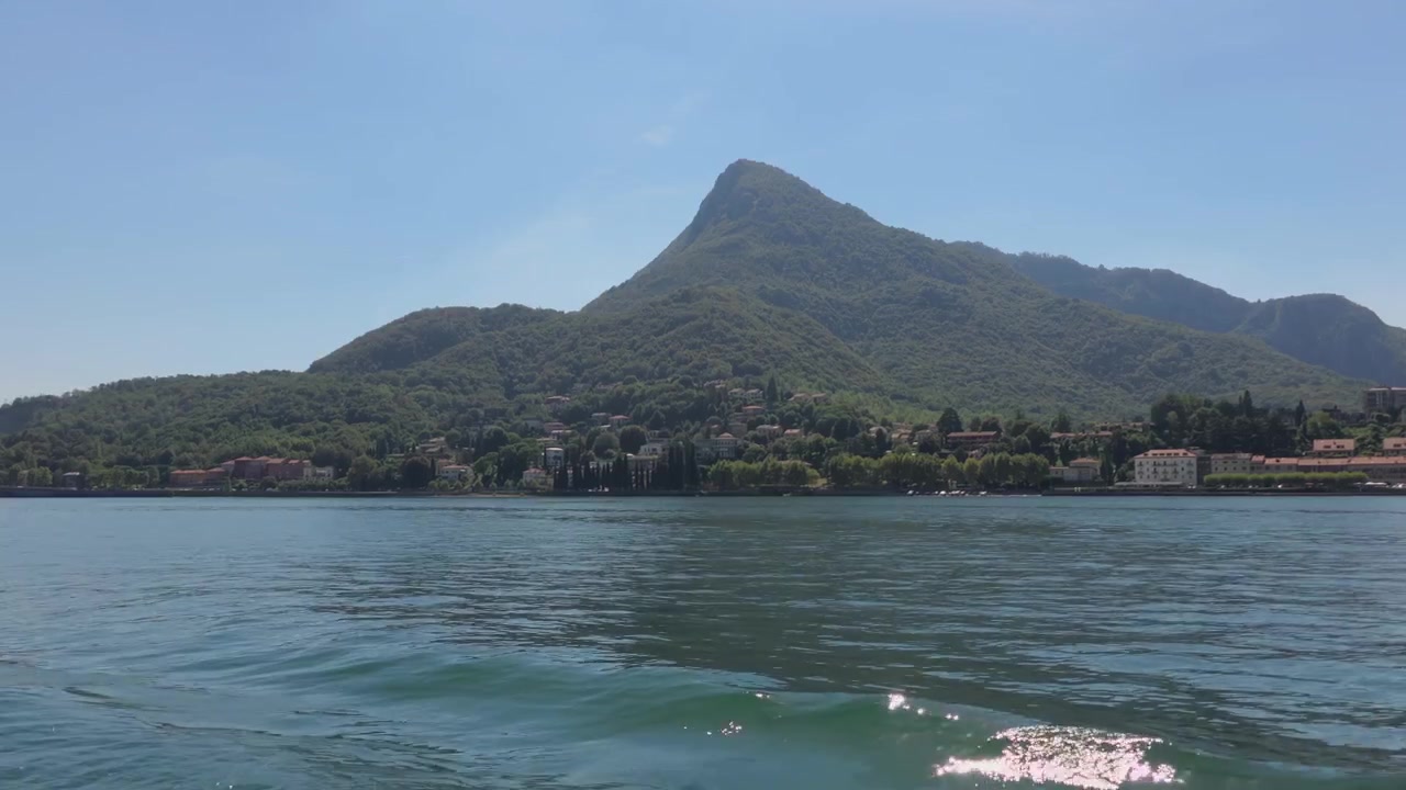 Ripresa panoramica serena del Lago di Lecco con acque blu calme, montagne verdeggianti e pittoreschi edifici in riva al lago sotto un cielo limpido e soleggiato