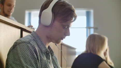 a male student in a university auditorium listens to music in white headphones during a break without paying attention to others. a lot of people in the audience are talking.