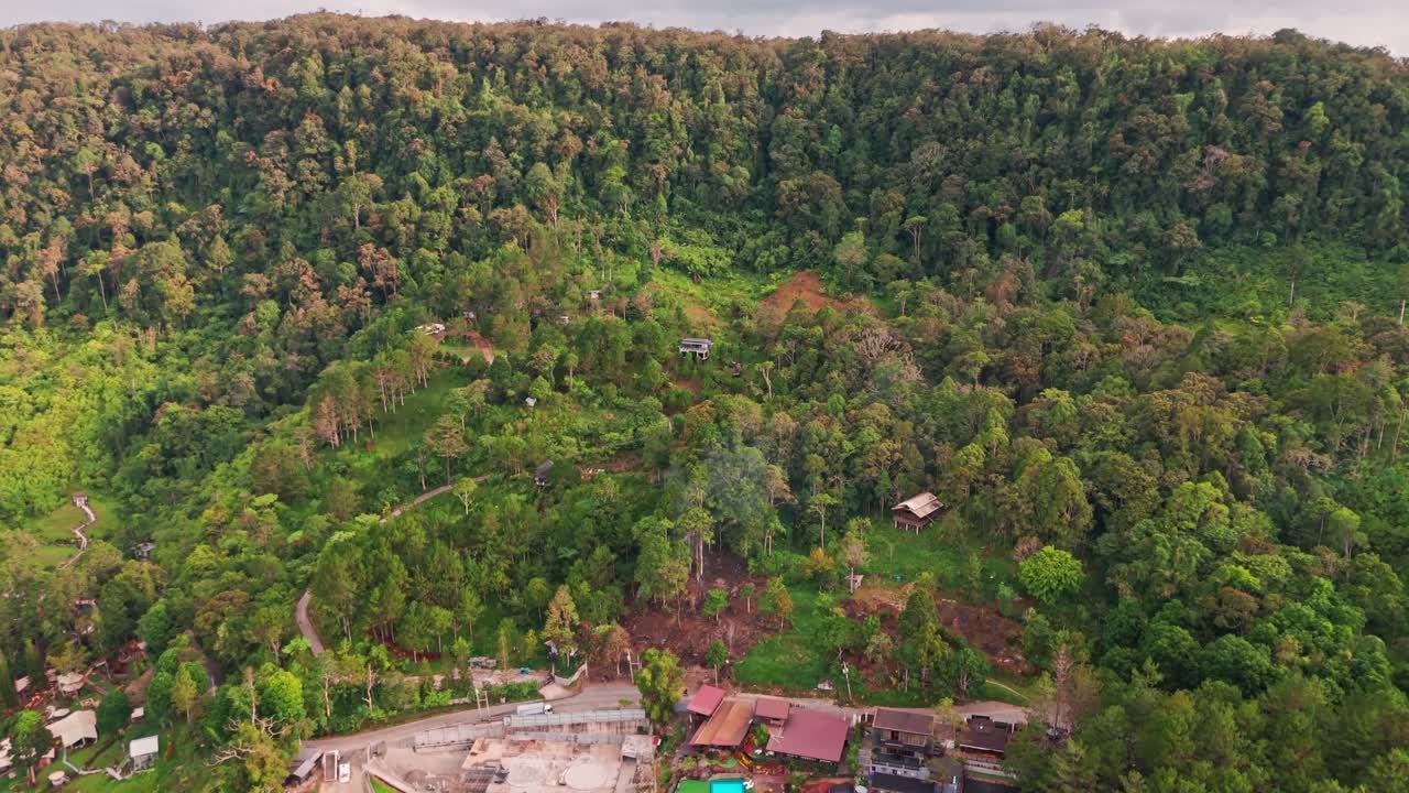 Aerial drone view of dense tropical forest at golden hour, revealing lush canopy, hillside homes, warm sunlight, and dramatic mountain clouds across a vibrant natural landscape
