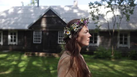 a woman with the flower crown is walking by an ancient cottage as the camera is following her, natural colour, slow motion