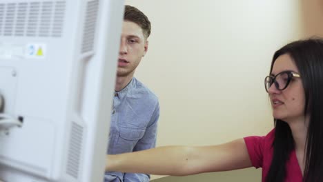 Young-man-talking-to-attractive-female-doctor,-physician-prescribing-medication-to-the-patient-and-explaining-the-results-of-ultrasonography-pointing-at-the-screen-of-the-computer-in-clinic