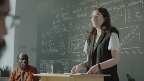 woman giving a scientific lecture in a classroom with equations on a chalkboard