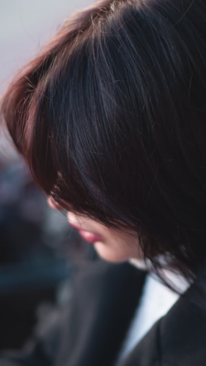 부드럽고 흐르는 갈색 머리카락과 사려 깊은 표정을 가진 여성의 클로즈업, 야외 도시 환경, 배경은 흐릿한 식물과 미묘한 밝은 톤을 특징으로합니다.