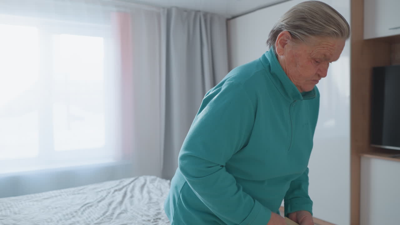 Elderly woman in teal sweater walking away from bed towards the window, calm home environment with soft natural light, minimalist room design, serene indoor atmosphere, bright curtains and simple decor