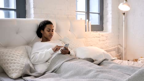 young african girl using smartphone while relaxing in bed