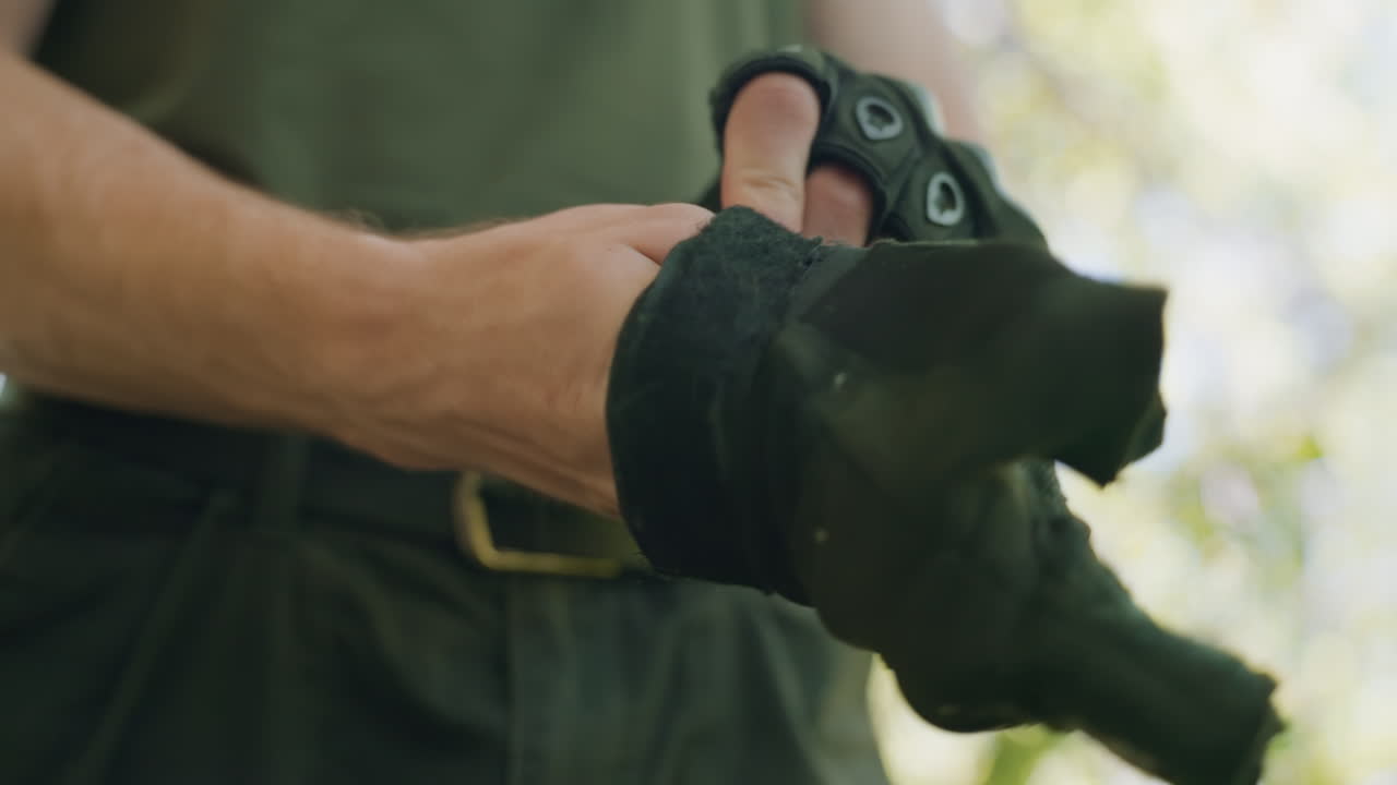 Close up view of soldier adjusting fingerless glove strap around wrist, veins visible on forearm, black cargo pants and belt shown, blurred green foliage and sunlit forest floor backdrop