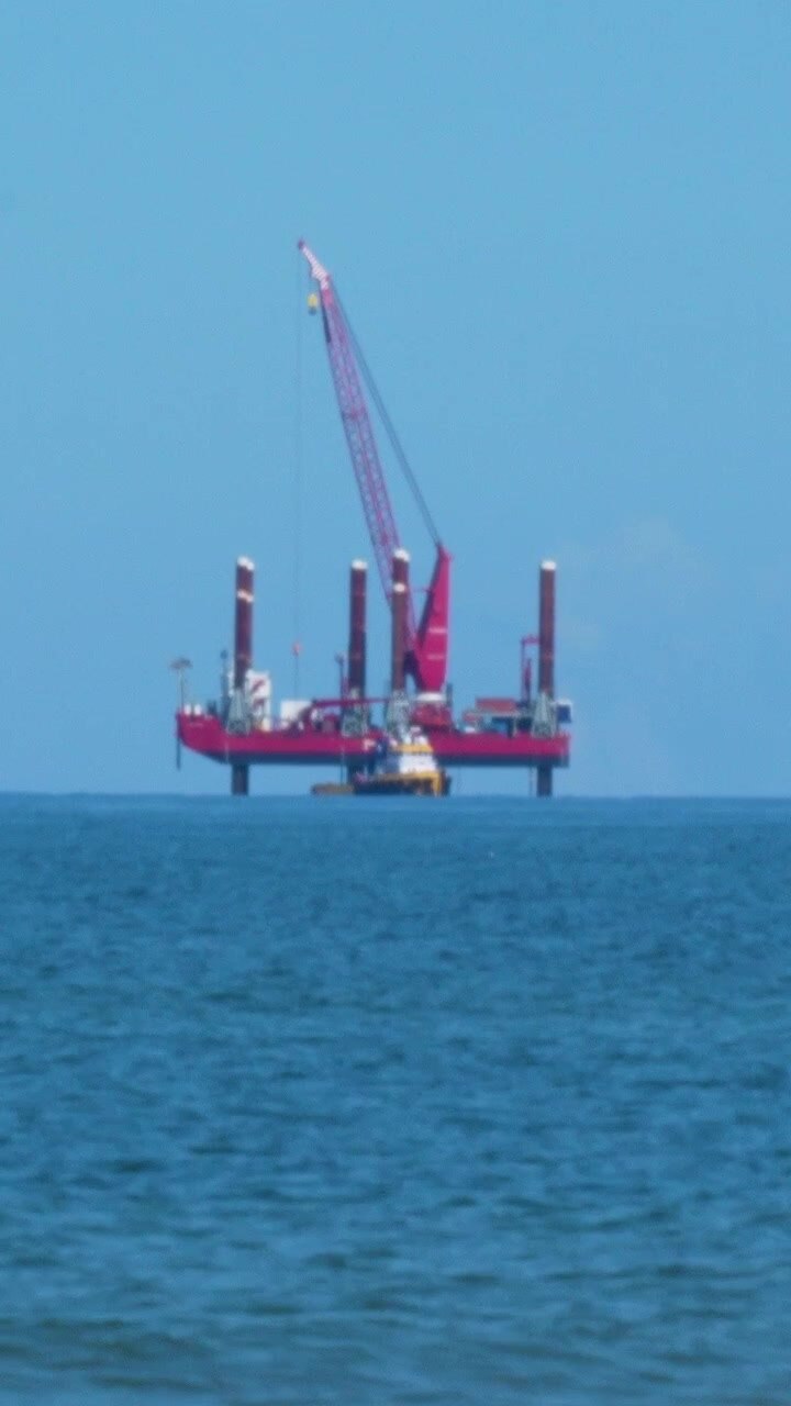 A red jackup rig or offshore construction vessel with a large crane stands in the blue waters of the Baltic Sea near Osetnik, Poland, representing energy and industrial infrastructure.