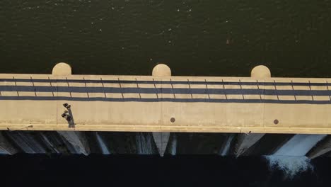 Ascending-top-down-shot-of-couple-walking-on-bridge-dam-over-river-during-sunny-day