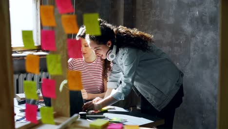 female designer is working with laptop when her colleague is entering office. she is listening to misic, then taking off headphones and start helping her coworker.