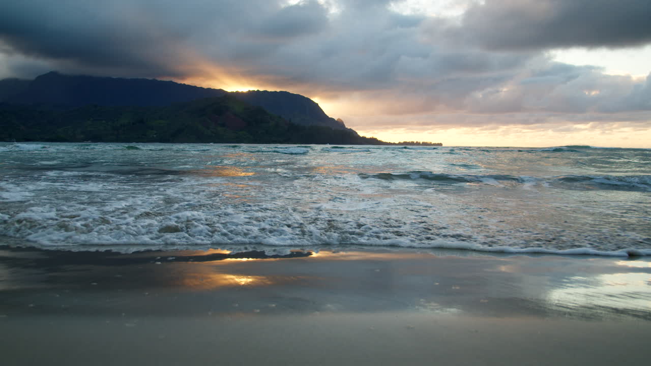 un primer plano de las olas rodando sobre la arena reflectante de hanalei en un atardecer temperamental en cámara lenta