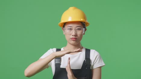 close up of asian woman worker wearing goggles and safety helmet looking at camera and showing time out hands gesture while standing in the green screen background studio