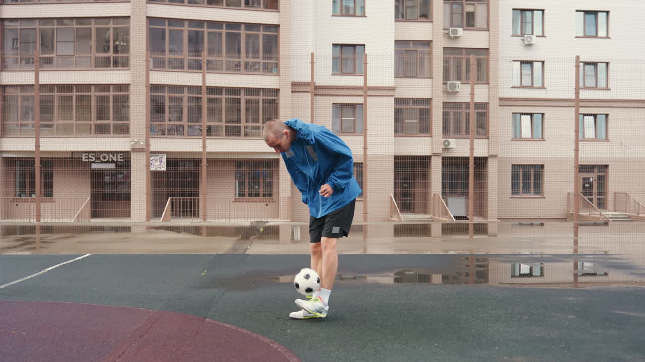 mavi ceket ve spor ayakkabı giyen bir adam, yağmur sırasında şehirdeki bir yerleşim bölgesinin oyun alanında ıslak zeminde futbol topu sürüyor, su birikintileri apartman cephelerini yansıtıyor, ritmik ayak hareketleriyle tek başına antrenman seansı