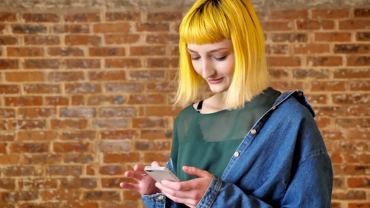 Giovane adolescente insolito con i capelli gialli in piedi che messaggia al telefono vicino a un muro di mattoni sullo sfondo, sorridente e positivo.