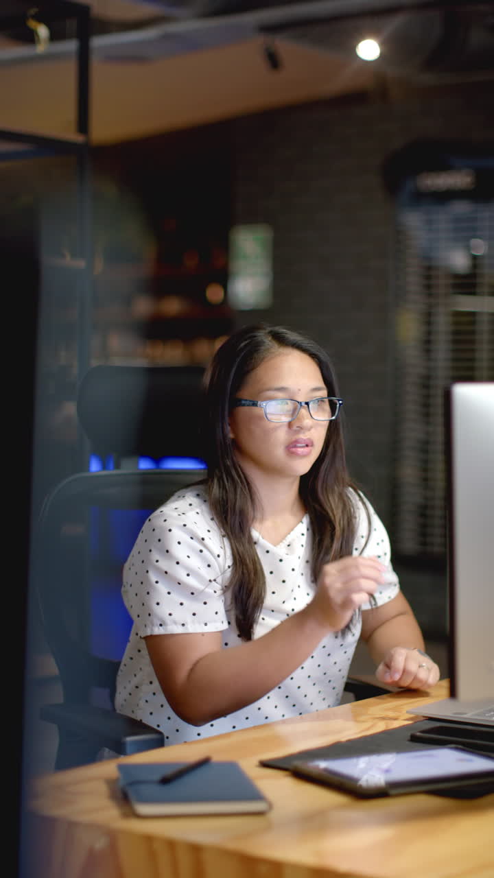 Vertical video: Working late, female business professional thinking while holding glasses at desk