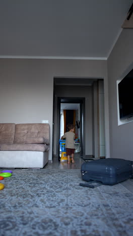 lovely toddler pushes a toy cart with a big yellow ball inside. kid walks by the big suitcase lying on the floor in the room. vertical video.