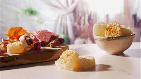 a hand gently lifts a cube of turkish delight topped with crispy golden kataifi dough from a small stack, as warm sunlight highlights a rustic board of assorted sweets and rosebuds in the background.