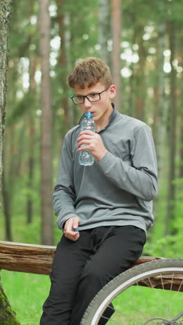 un niño joven con gafas y una camiseta de manga larga gris abre una botella de agua y toma un sorbo mientras está sentado en un tronco en el bosque, descansando al lado de una bicicleta estacionada rodeada de altos árboles verdes