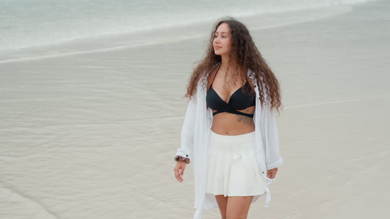 Modella in costume da bagno che passeggia sulla sabbia bagnata. Una donna elegante in abito estivo cammina su una spiaggia costiera. Una donna alla moda che avanza con sicurezza sulla spiaggia con piccole onde e uno sfondo soleggiato.
