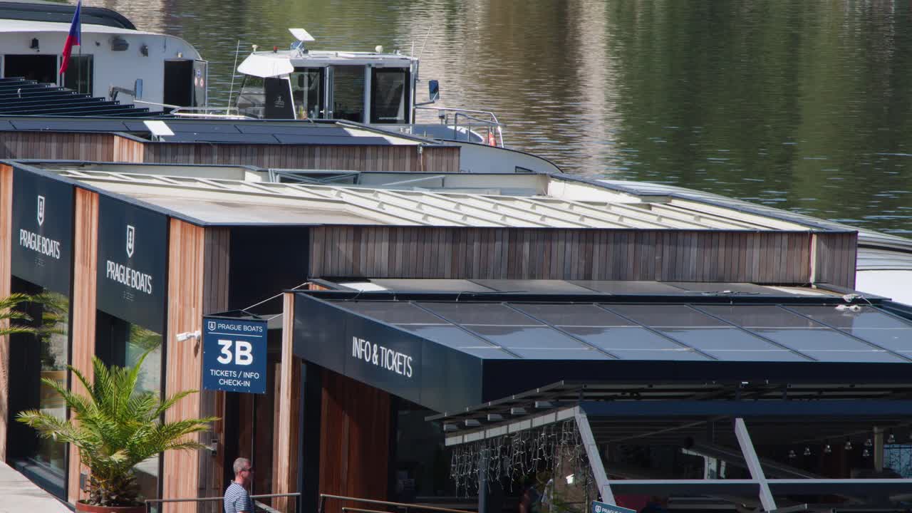 Man walks toward modern riverside ticket booth, boats docked, sunny summer day, Prague, Czech Republic