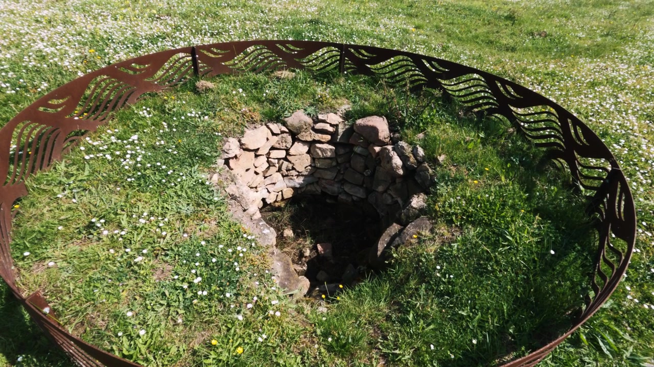 Ruined stone well surrounded by green grass and wildflowers, encircled by a decorative rust metal ring.
