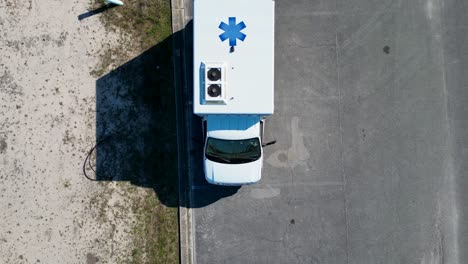 Vista-Aérea-Que-Se-Eleva-Lentamente-De-La-Ambulancia-En-El-Estacionamiento-Al-Lado-Del-Lote-De-Arena