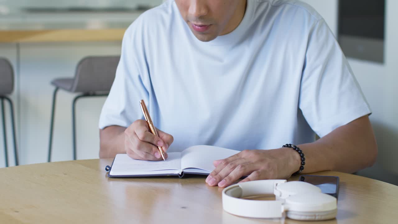Adult man reaching for metallic pen and writing in notebook at kitchen table, studying notes