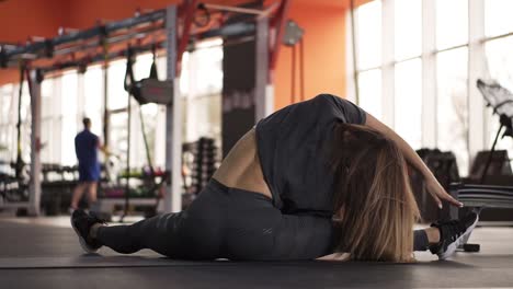 Footage-of-muscular-back-of-an-athletic-slim-woman-who-practices-fitness-and-sits-in-a-modern,-sportive-gym.-She-stretches-her-legs-and-bends-to-her-leg-actively.-Slow-motion