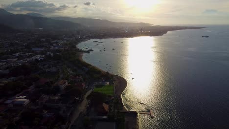 drones aéreos bajando sobre la ciudad capital de dili, timor leste en el sudeste de asia con hermosa luz solar dorada reflejándose en el agua del océano y barcos amarrados en aguas tranquilas a lo largo de la costa