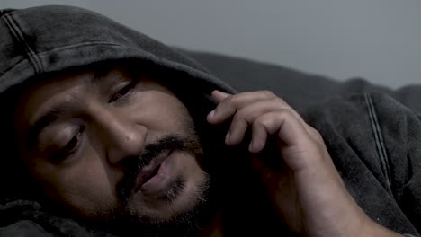 Close-up-shot-of-the-face-of-an-Indian-man-wearing-a-hoodie-half-asleep-talking-on-his-mobile-phone,-receiving-a-late-night-call-from-his-staff-as-he-is-the-duty-manager-for-after-hour-emergencies