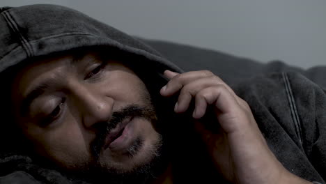 close up shot of the face of an indian man wearing a hoodie half asleep talking on his mobile phone, receiving a late night call from his staff as he is the duty manager for after hour emergencies