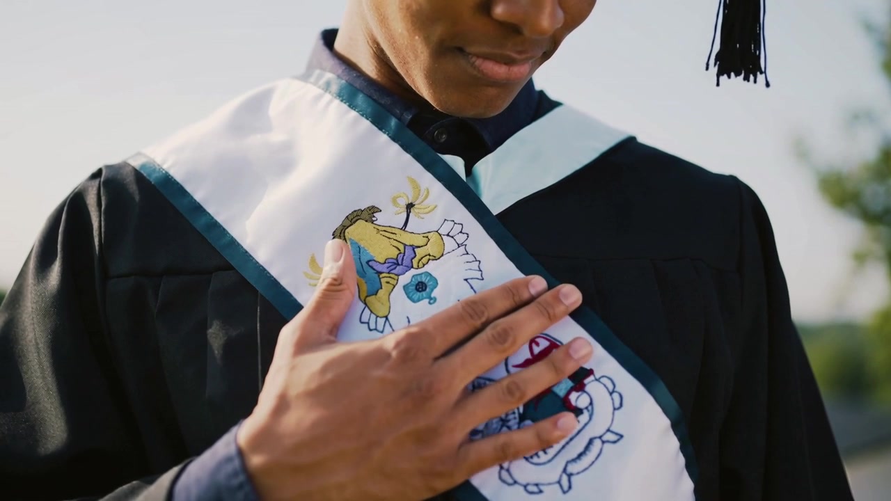 Looking down graduate in black gown adjusting white sash at rooftop, revealing crests and tassel