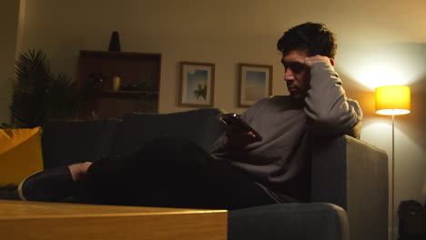 young man spending evening at home sitting on sofa with mobile phone scrolling through internet or social media 9