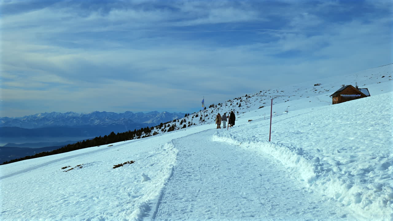 Family hiking on a snowy mountain path near a Bavarian flag and a wooden cabin under a clear sky.