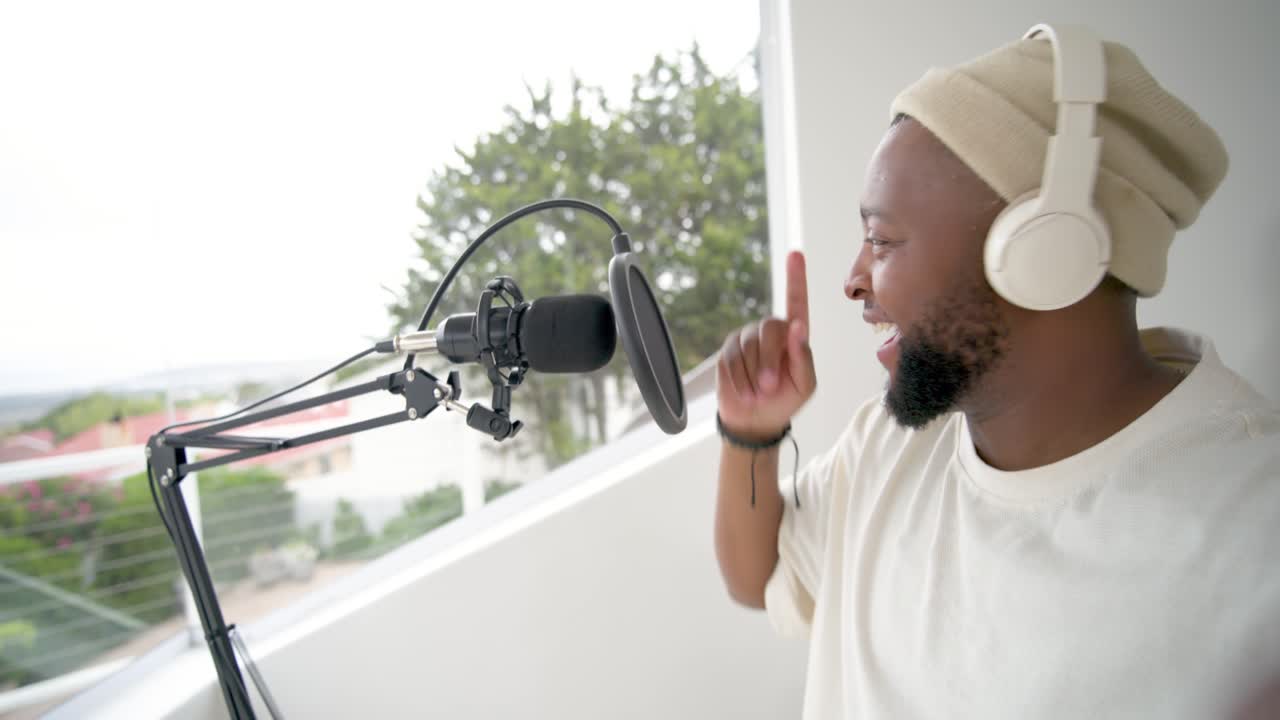 Adult African man recording at home studio, tapping camera, leaning into mic, pointing to show view