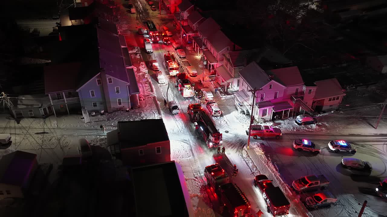 Aerial night view of snow-covered American neighborhood with police cars and fire trucks responding to an emergency, flashing lights illuminating icy streets during coordinated operation. Approaching