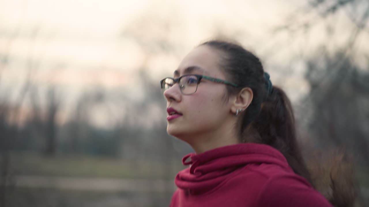 Woman Jogging Peacefully, Evening Run With Reflective Mood, Profile Shot Of Woman With Glasses During Evening Exercise, Silhouetted Woman Wearing Hoodie And Glasses Pauses Amidst Trees At Sunset