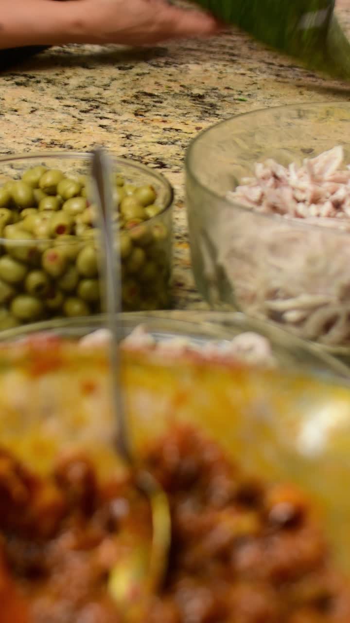 Vertical video. Ingredients in focus while a woman assembles a traditional hallaca on a banana leaf in the background during food preparation