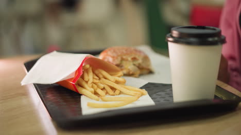 a close-up of fries, burger, and coffee cup on a tray, the woman s hand reaches for fries, takes a bite, and sips her coffee, all while a soft bokeh lighting effect beeps in the blur background