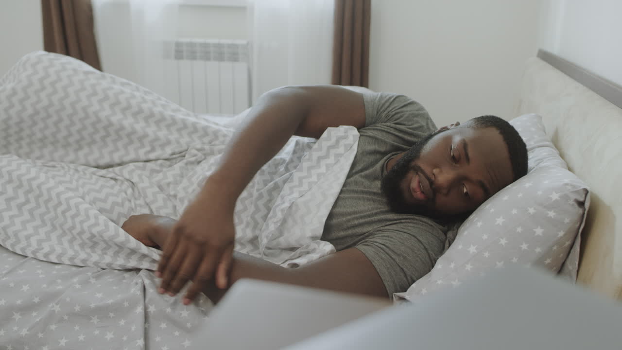Uomo assonnato che si alza dal letto in camera. Giovane che prende la sveglia in mano.