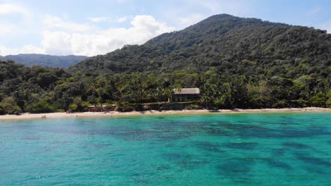 Aerial-view-over-shallow,-turquoise-sea,-overlooking-a-beach-and-jungle-on-the-coast-of-Tayrona-national-natural-park,-sunny-day,-in-Colombia---tracking,-drone-shot