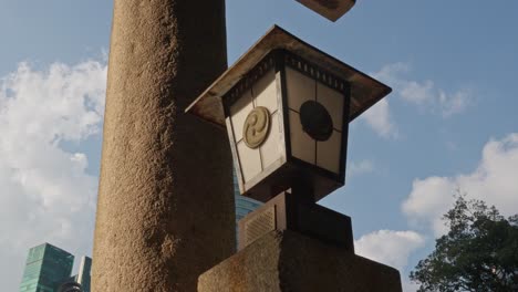 a weathered stone column and traditional japanese lantern with a distinctive symbol stand against a backdrop of modern high-rise buildings and a cloudy sky.