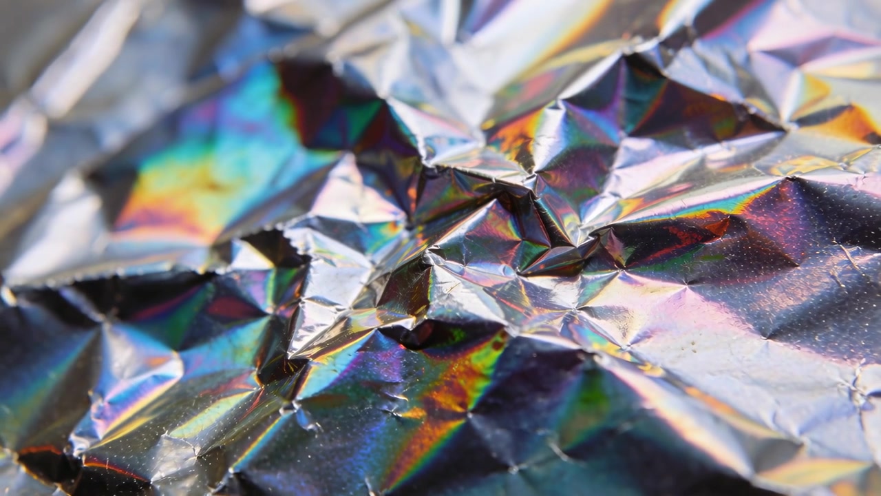 Telecamera macro che si sposta mettendo a fuoco sopra un tavolo per rivelare una pellicola iridescente accartocciata con riflessi arcobaleno
