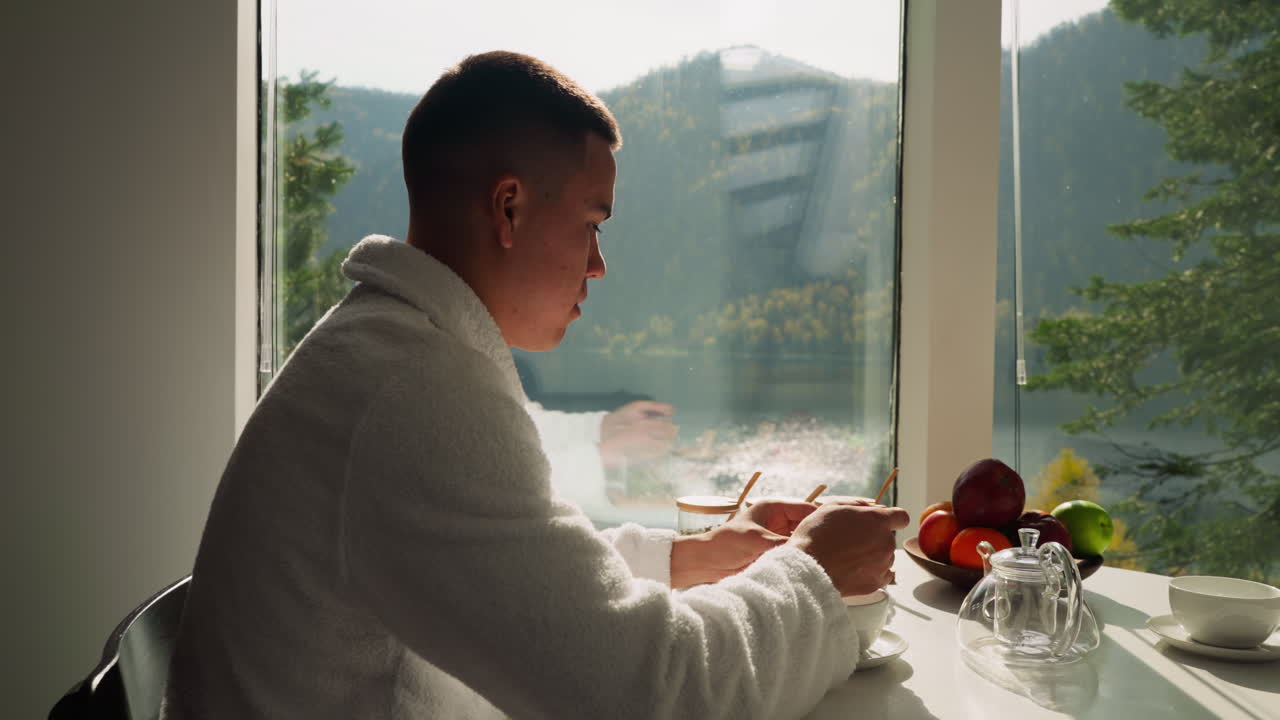 한 남자가 창문 에 앉아 차를 준비하고 있다. 집안 옷을 입은 집중된 남자가 주방에서 술을 마시기 위해 항아리에서 건조한 잎을 가져간다. 아침 의식