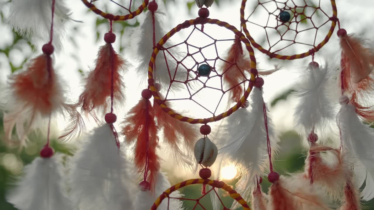 cazador de sueños con plumas naranjas balanceándose en cámara lenta deslizándose con una ligera brisa en el árbol en el bosque en la luz del atardecer en primavera en un día soleado. amuleto de estilo boho colgando en un árbol en el parque contra el cielo azul