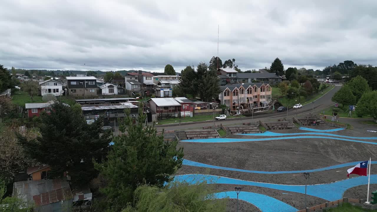 Volo aereo all'indietro che rivela una piccola piazza cittadina con strutture parasole blu, circondata da case residenziali e colline verdi sotto un cielo coperto. Entre Lagos, Puyehue, Cile.