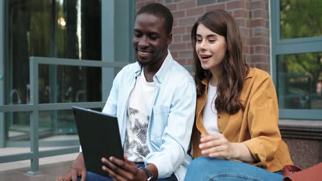 hombre afroamericano y mujer caucásica sentados en la calle y haciendo una videollamada en una tableta cerca de la universidad