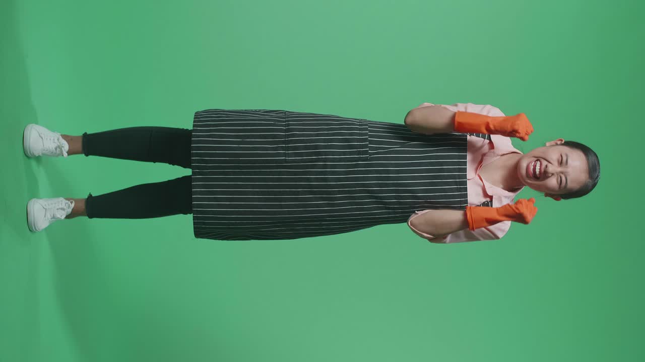 cuerpo lleno de feliz ama de casa asiática con un delantal celebrando mientras estaba de pie en el estudio de pantalla verde