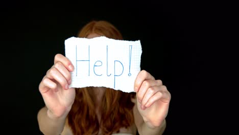 Young-redhead-woman-holding-paper-card-with-help-inscription-on-black-background