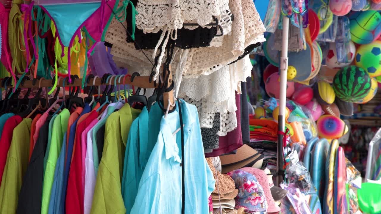 Un'inquadratura media statica cattura vivaci capi d'abbigliamento estivo e bikini appesi in un mercato thailandese